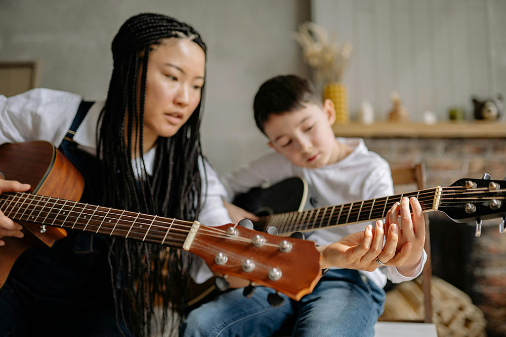 A woman with long braids guides a young boy's hand on a guitar neck. Guitar lessons Auckland